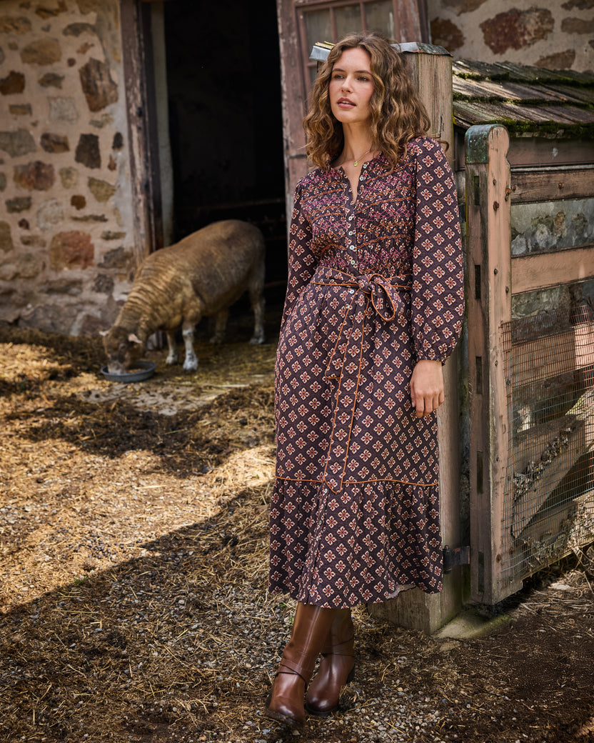 Model wearing the Desert Primrose Shay Dress, showing its patterned print, long sleeves, waist tie, and tiered skirt near a farm outbuilding.