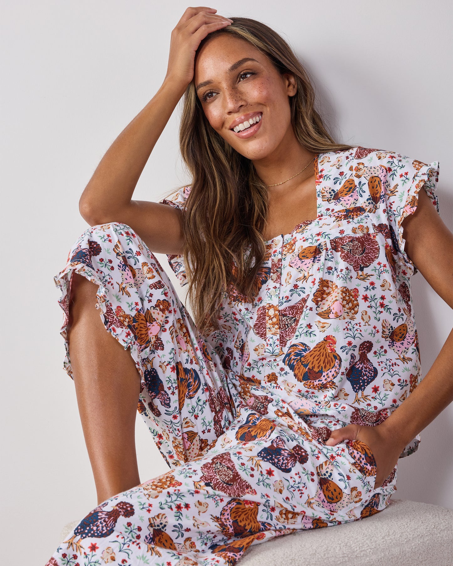 Model seated wearing a cropped pajama top with flutter sleeves and matching pants in a chicken-and-floral print.