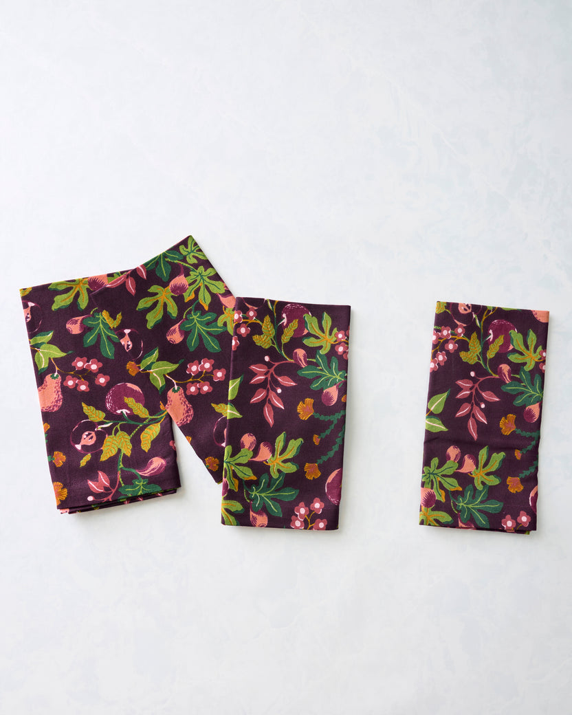 Folded Pomme & Poire dinner napkins in a plum fruit-and-floral print, arranged separately to show the pattern.