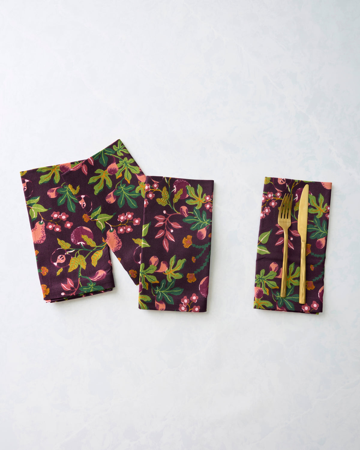 Folded Pomme & Poire dinner napkins in a plum fruit-and-floral print, with a fork and knife placed on one napkin.