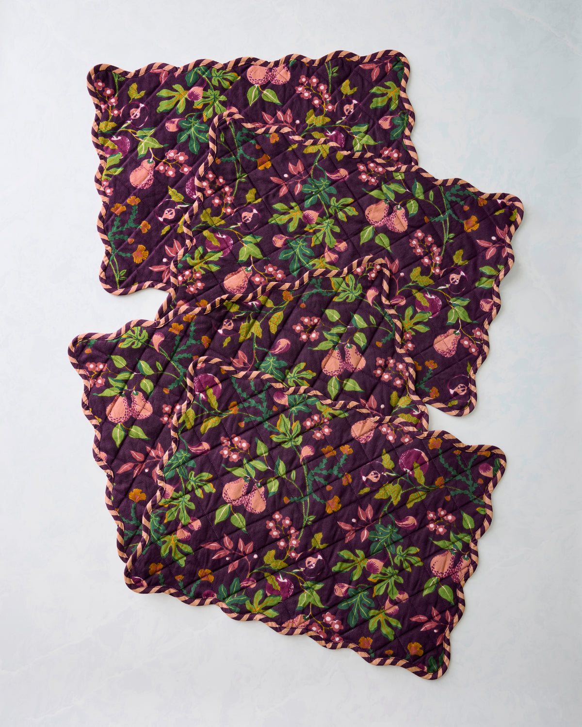 Four overlapping quilted Pomme & Poire placemats with scalloped edges and a plum fruit-and-floral print.