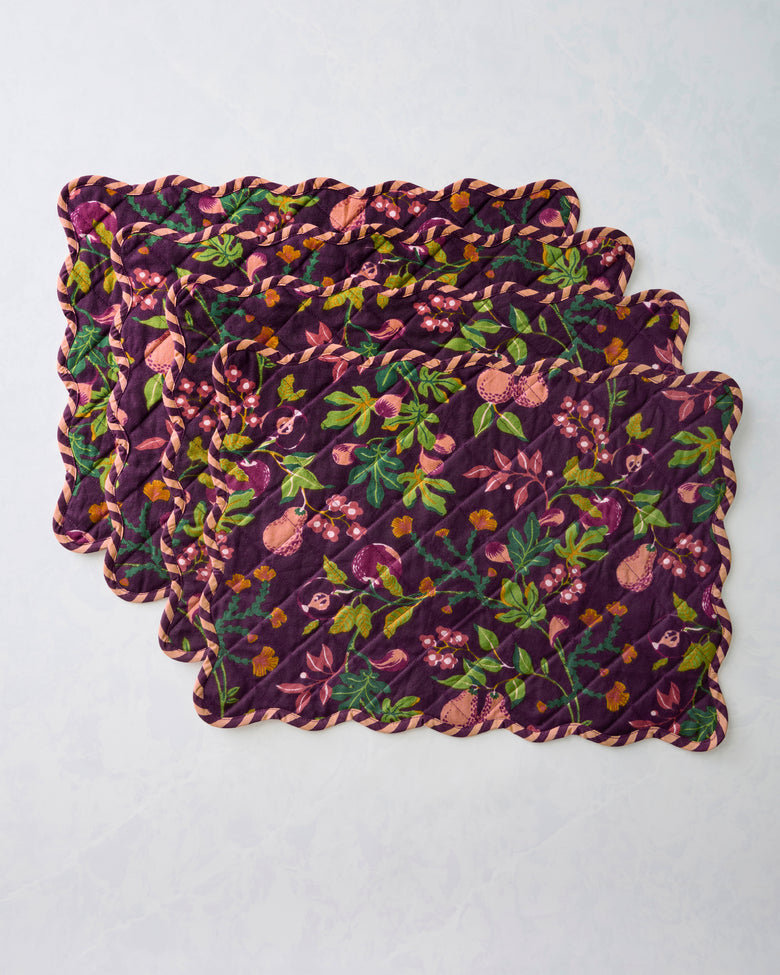 Four quilted Pomme & Poire placemats stacked with their scalloped edges and plum fruit-and-floral print visible.