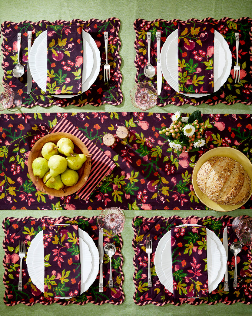 Styled dining table with four place settings using plum floral dinner napkins, matching placemats and a table runner, plus pears, bread, flowers, and glassware.