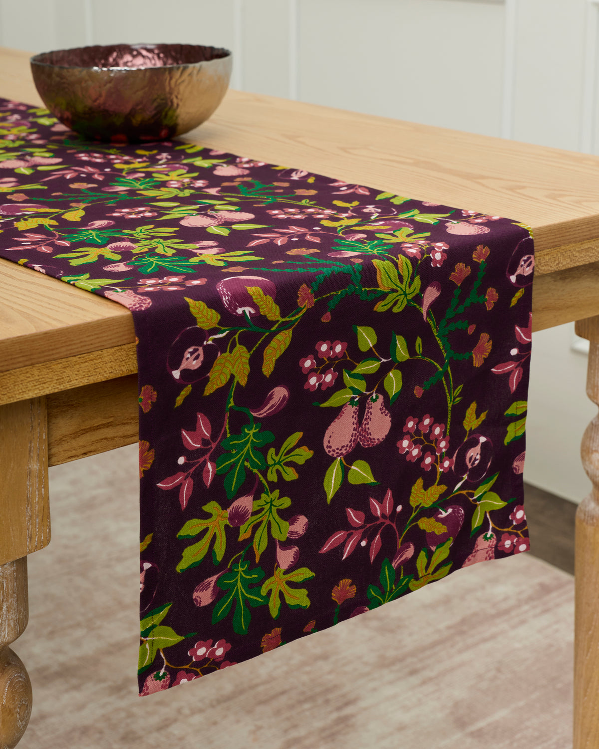 Plum Pomme and Poire table runner draped over a wooden table, printed with leaves, flowers, and fruit.