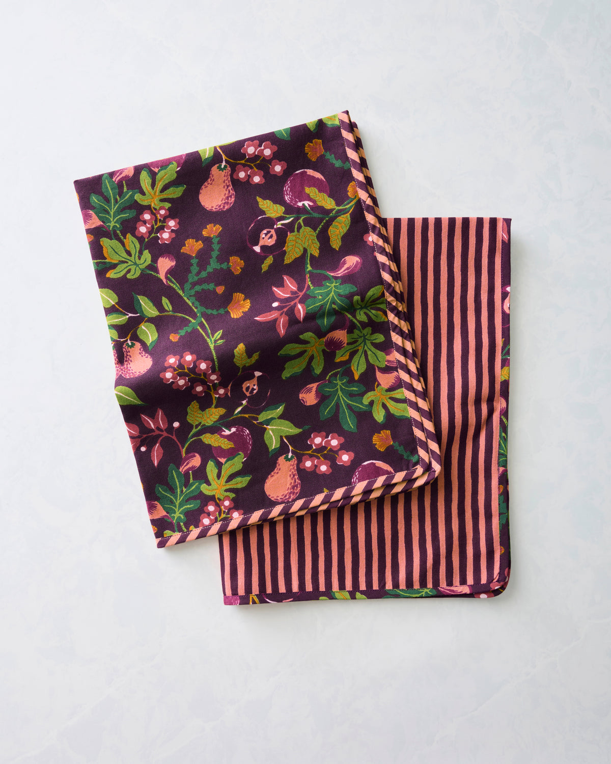 Two Pomme & Poire plum tea towels folded and overlapping, showing the fruit and floral print on one side and stripes on the other.