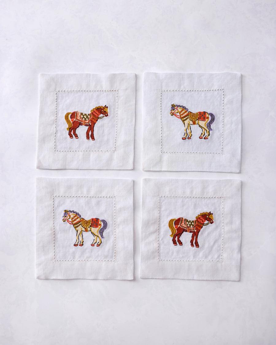 Set of four white cocktail napkins arranged in a square, each embroidered with a decorated horse.
