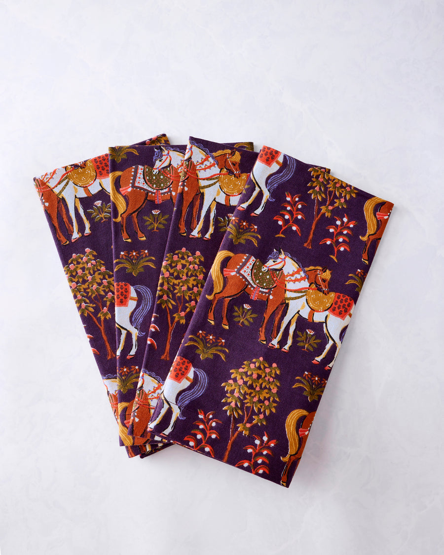 Four purple dinner napkins fanned out, printed with horses, trees, and floral motifs.