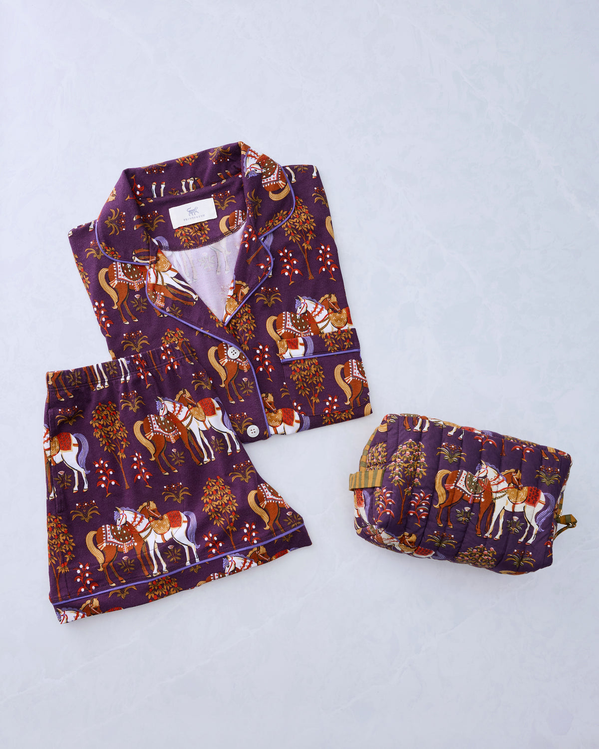 Deep aubergine short-sleeve button-front pajama set with horse print beside a matching quilted pouch.