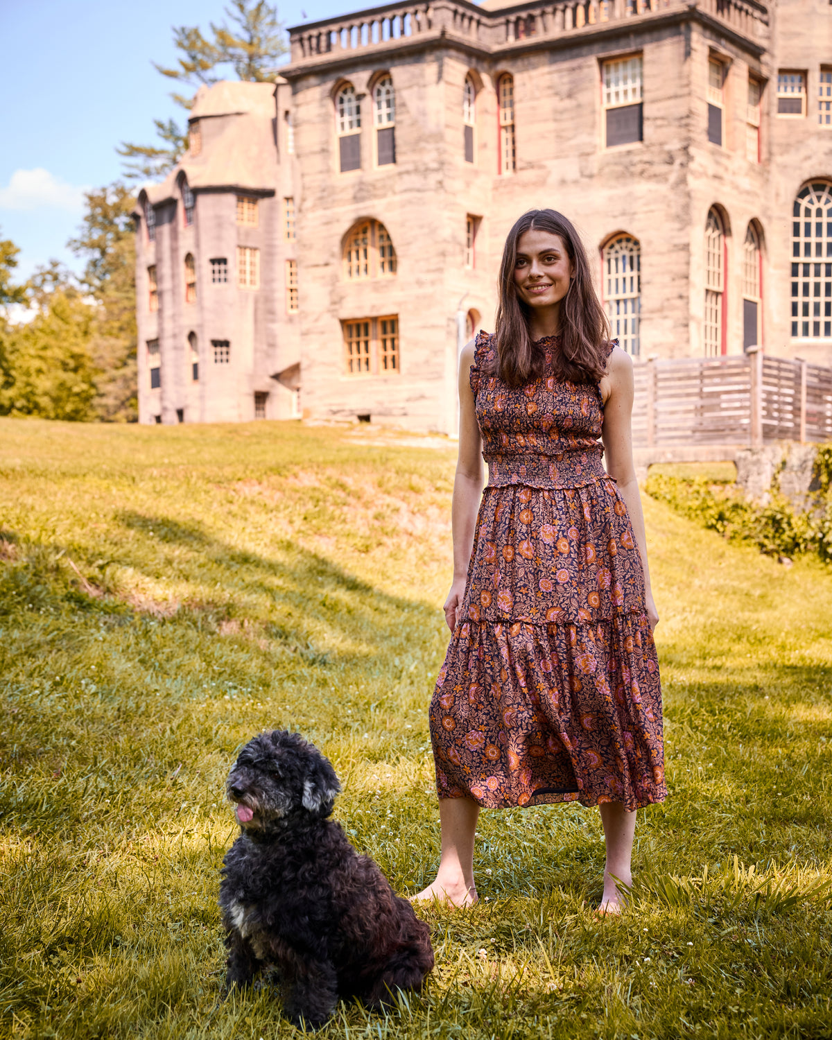 Model wearing the Sunset Vine Ellis Dress, a sleeveless patterned midi dress, on a lawn with a dog and large stone building behind her.