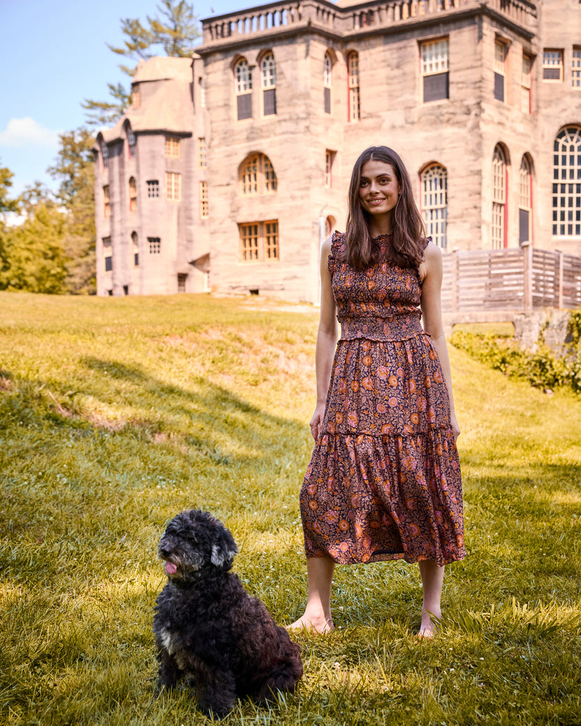 Model wearing the Sunset Vine Ellis Dress, a sleeveless patterned midi dress, on a lawn with a dog and large stone building behind her.