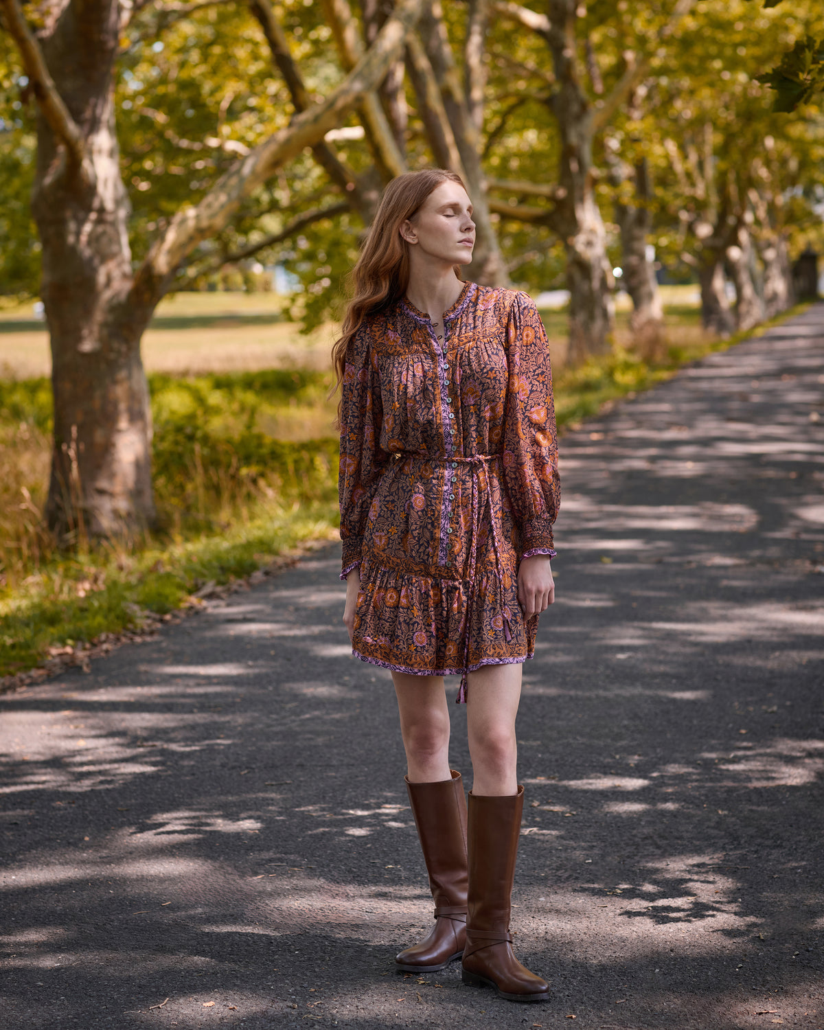 Front view of the Sunset Vine Soleil Dress, showing its floral print, long sleeves, button front, tie waist, and tiered skirt, worn with tall brown boots.