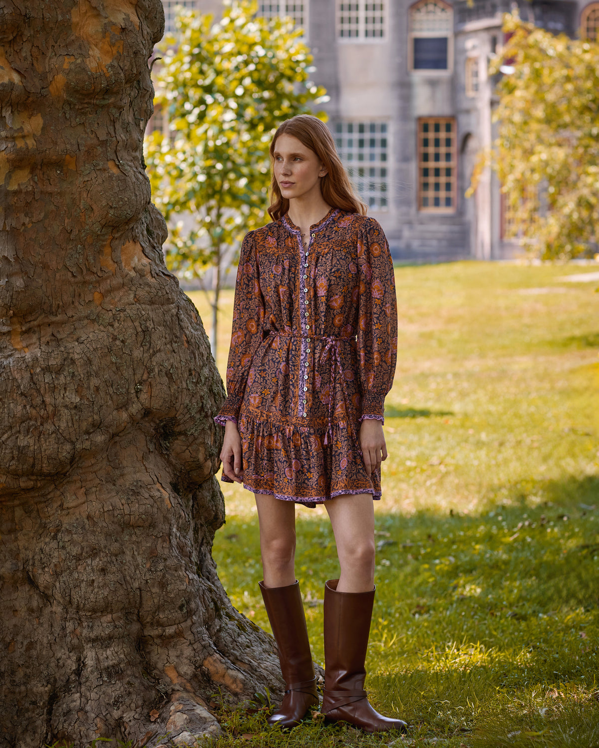 Front view of the Sunset Vine Soleil Dress, showing its long sleeves, button front, tie waist, and tiered skirt beside a large tree.