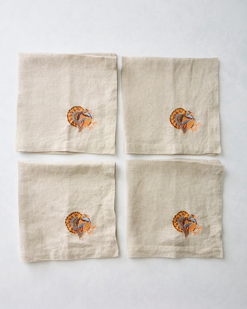 Four natural dinner napkins arranged in a two-by-two layout, each embroidered with a turkey.