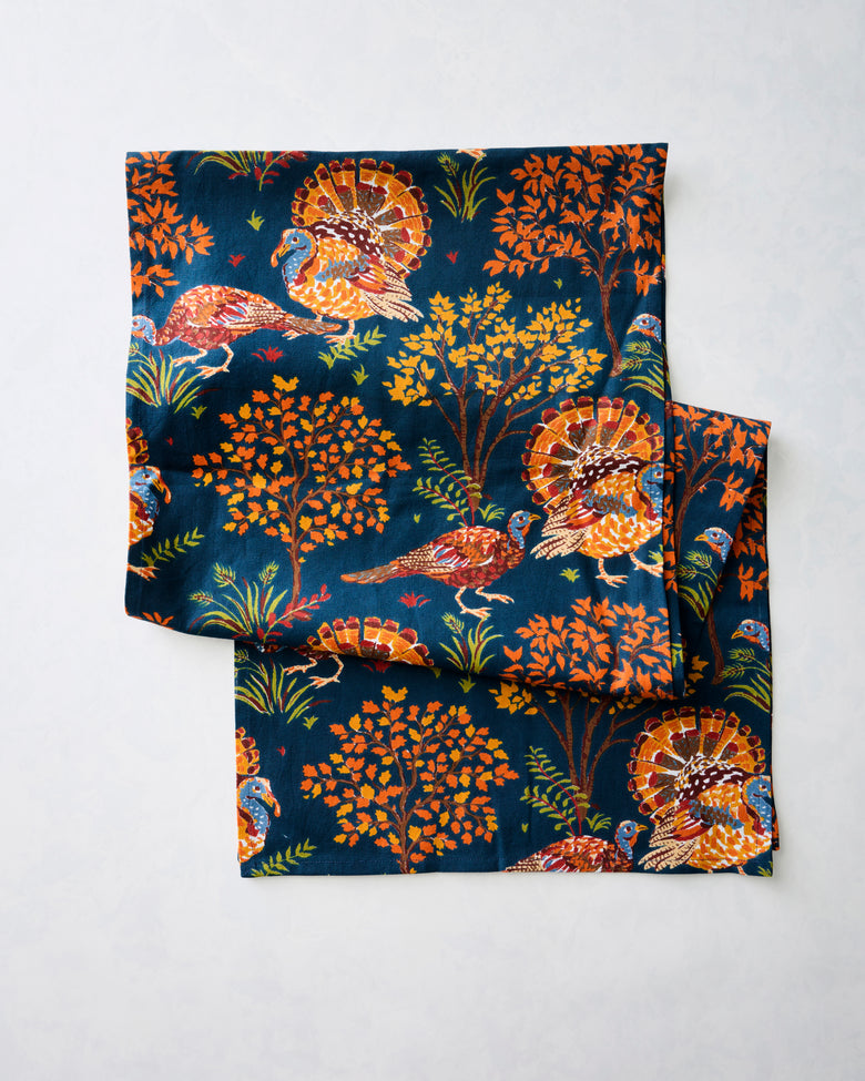 Folded navy table runner printed with colorful turkeys, trees, grasses, and leaves.