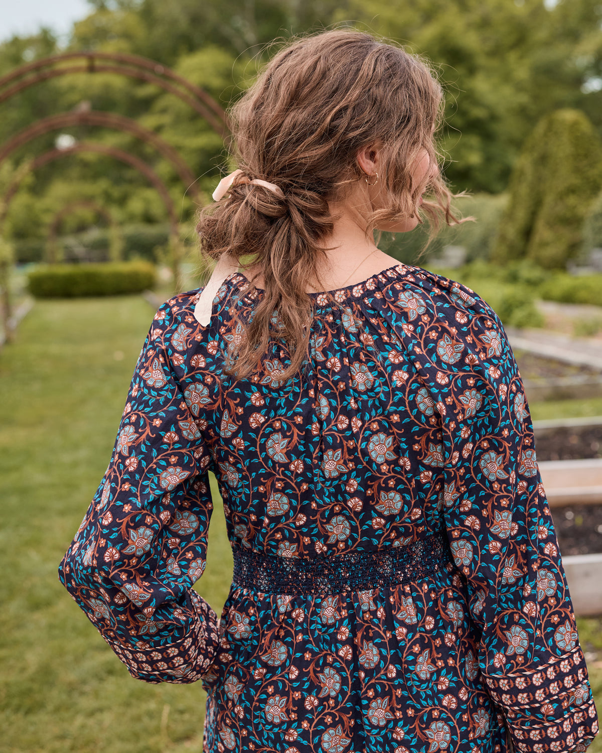 Back view of the Midnight Sky Flora Dress showing its full sleeves, patterned fabric, and smocked waist in a garden.