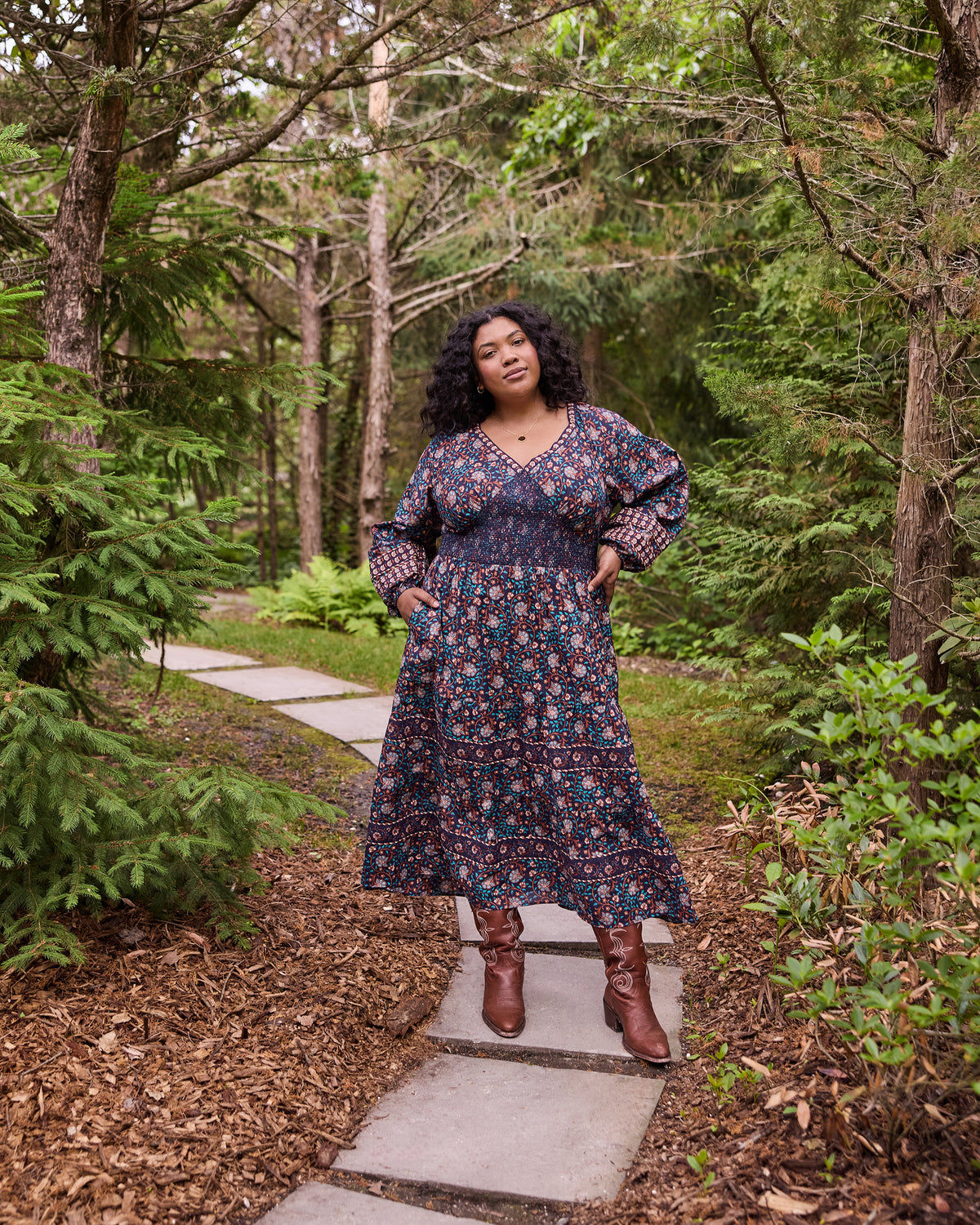 Front view of the Midnight Sky Flora Dress with a patterned print, smocked bodice, full sleeves, and pockets on a garden path.