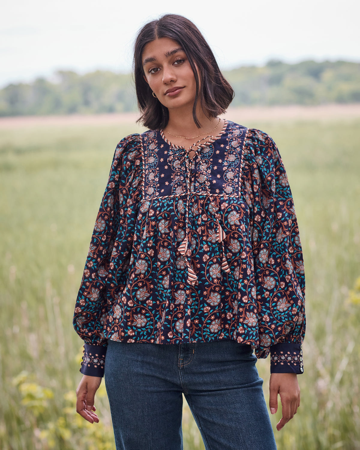 Front view of the Valley Garden Josephine blouse in a dark floral print, showing its billowy shape, criss-cross neckline, tassel ties, full sleeves, and button cuffs.
