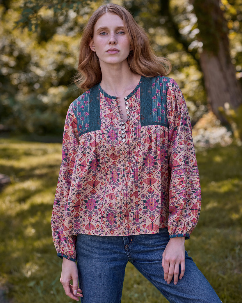 Model wearing a pink floral blouse with blue embroidered shoulder panels and jeans in a garden setting.