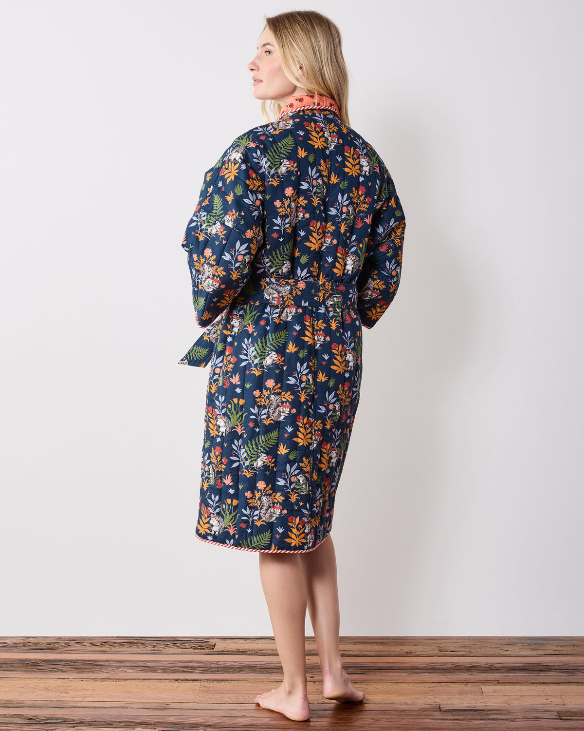 Back three-quarter view of an indigo quilted robe with a woodland animal and botanical print, matching belt, and striped hem trim.