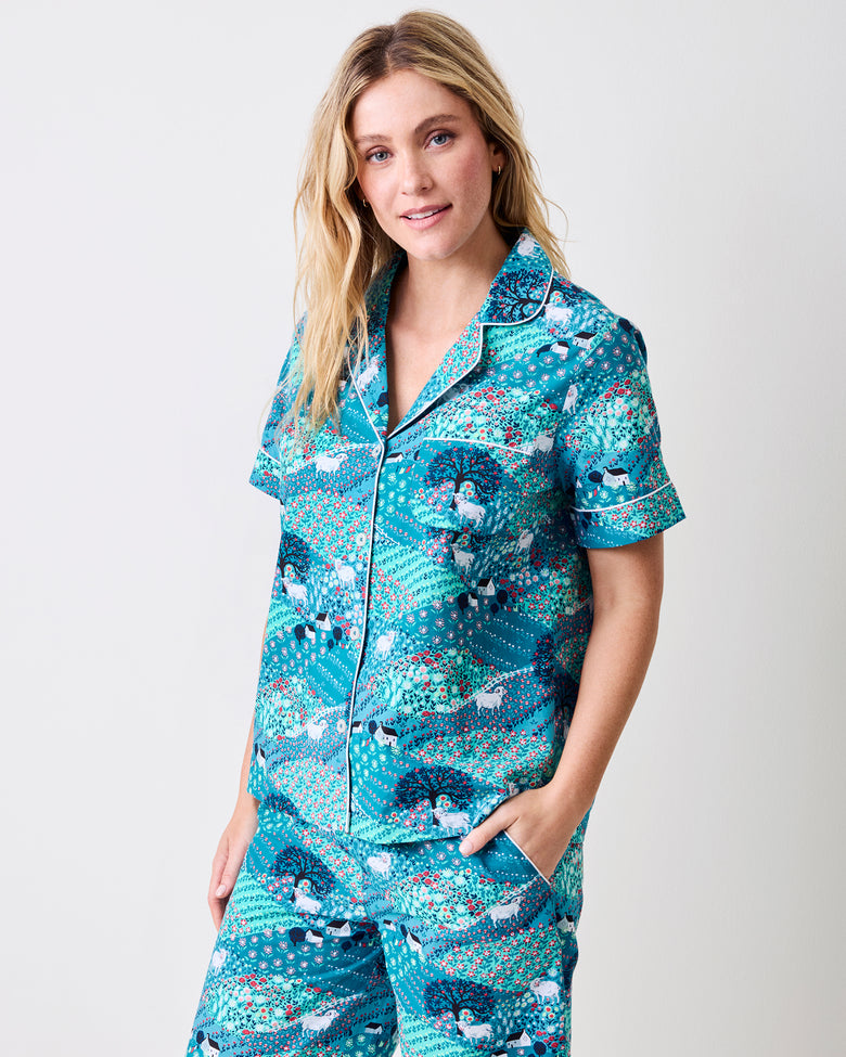 A woman modeling a matching short-sleeve button-front pajama top and pajama pants set with a teal, blue, and white peacock pattern, photographed against a light gray studio background.