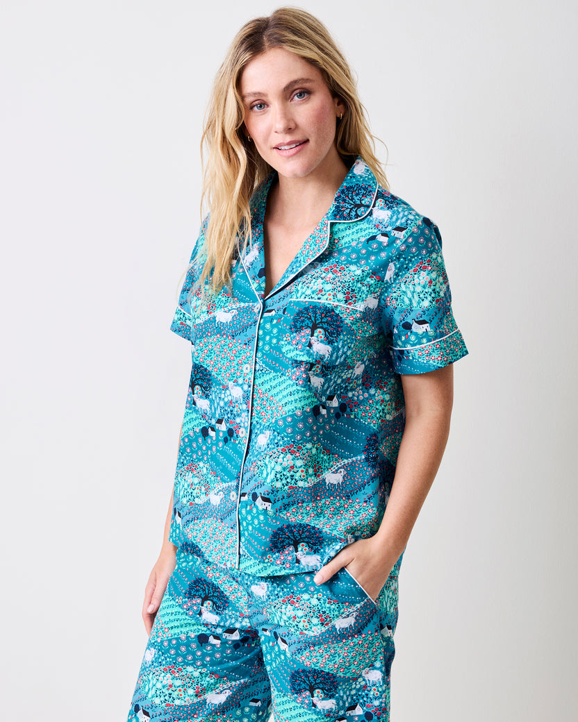 A woman modeling a matching short-sleeve button-front pajama top and pajama pants set with a teal, blue, and white peacock pattern, photographed against a light gray studio background.