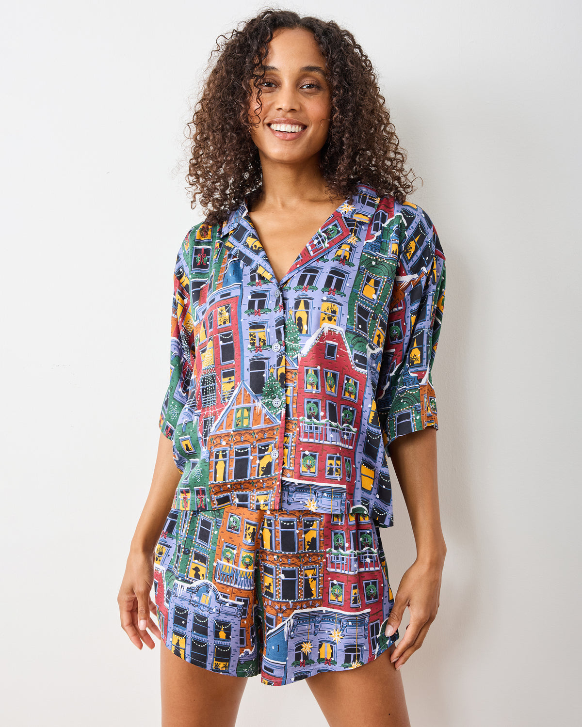 Front view of a person wearing the Manhattan Christmas Eco Satin Sweet Dreams set, featuring a patterned collared top and matching shorts with brownstone buildings and lit windows.