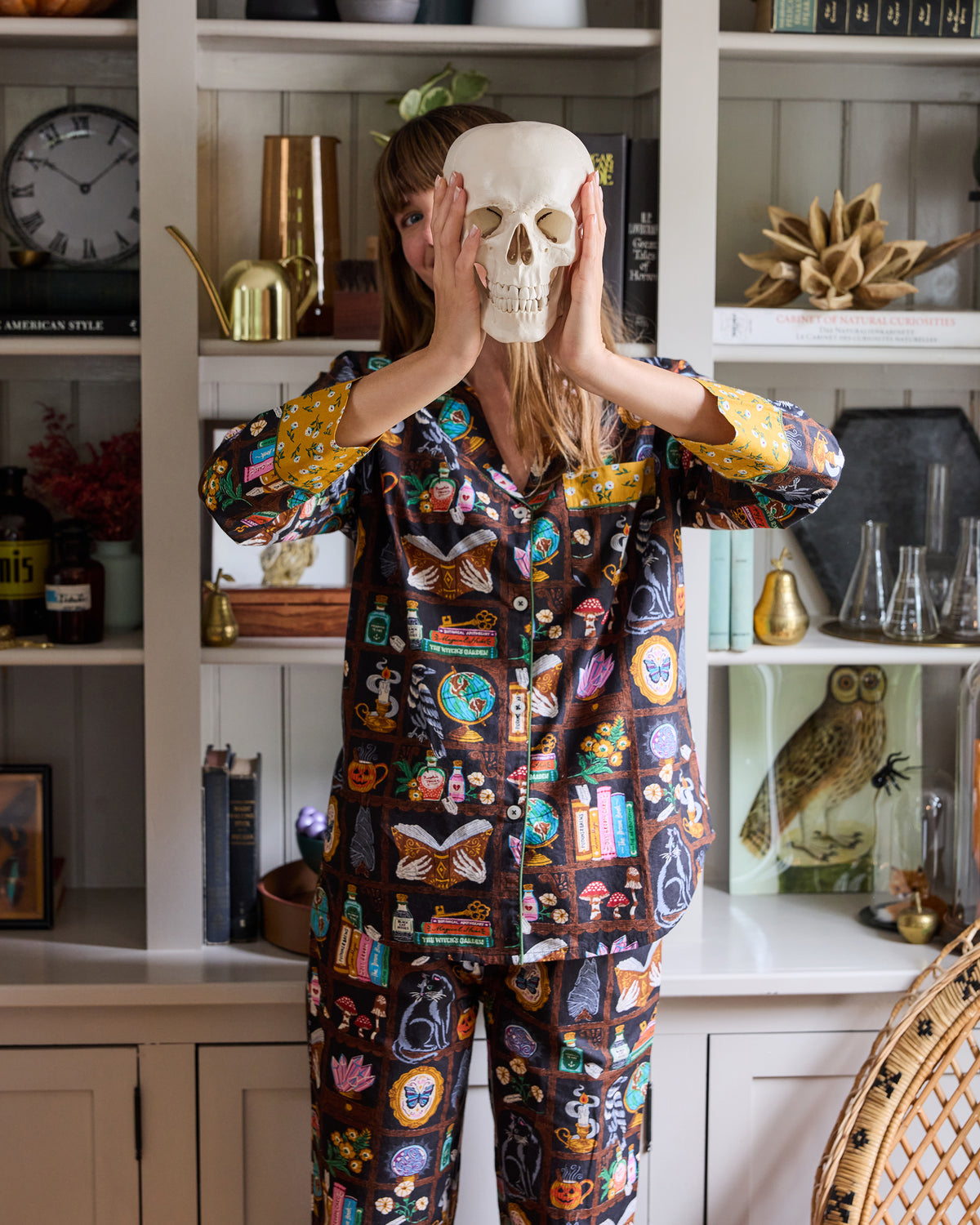 Front view of the Enchanted Study pajama set with a person holding a skull in front of the printed button-down top and long pants.