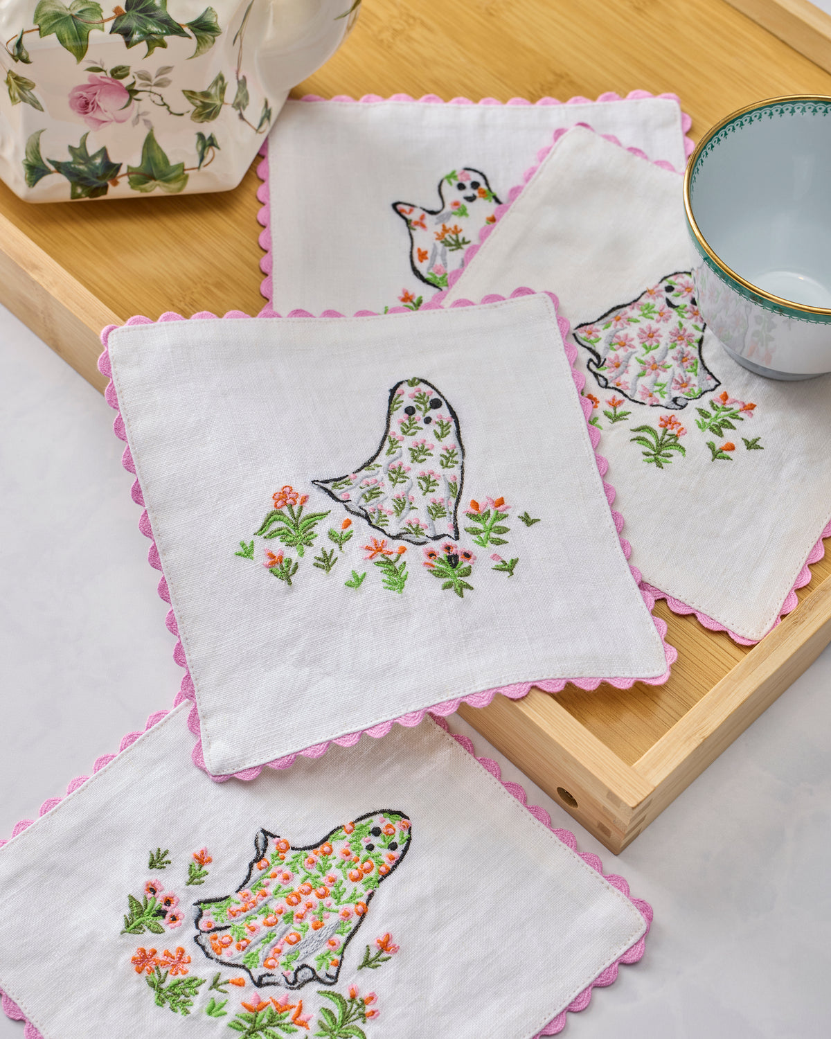 Gardening Ghosts embroidered cocktail napkins arranged on a wooden tray beside a floral container and teacup