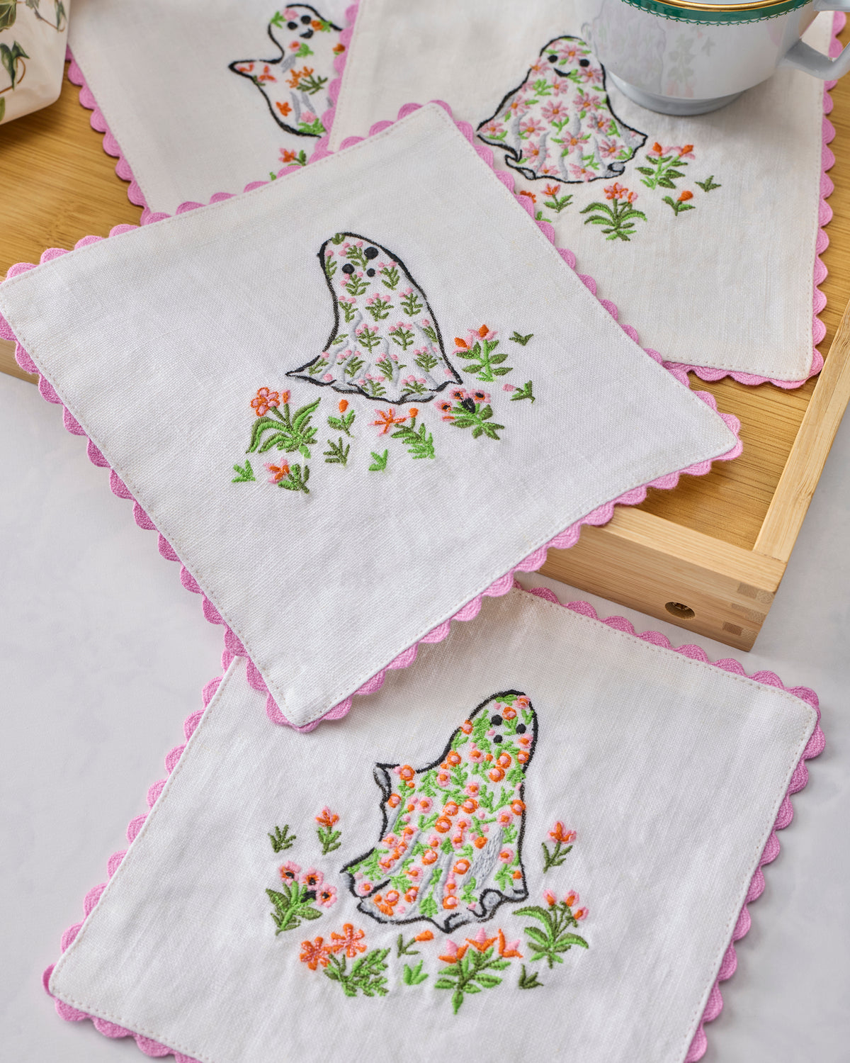 Four white cocktail napkins with pink scalloped edges, embroidered ghosts and flowers, arranged with a cup and tray.