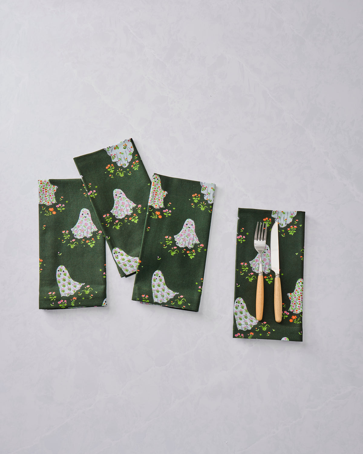 Three dark green dinner napkins with ghost and floral print, plus a folded napkin holding a fork and knife.