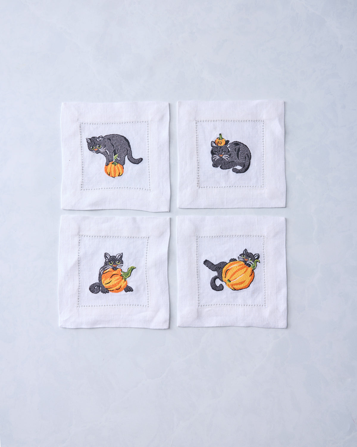Four white cocktail napkins arranged in a square, each embroidered with a black cat and orange pumpkin.