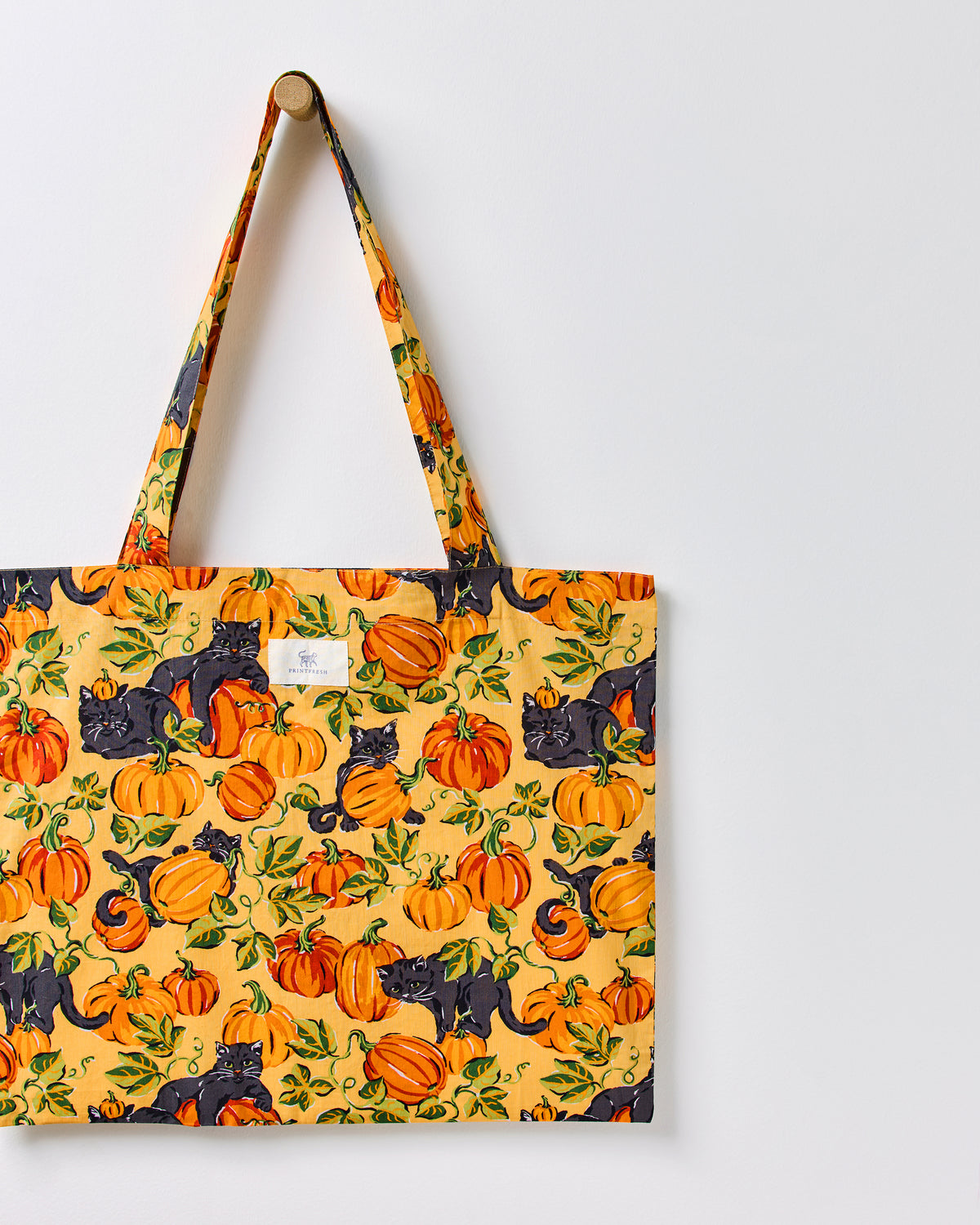 Close view of the Pumpkin Prowl cotton tote hanging from a wall peg, showing its orange pumpkin and black cat print, fabric handles, and center label.