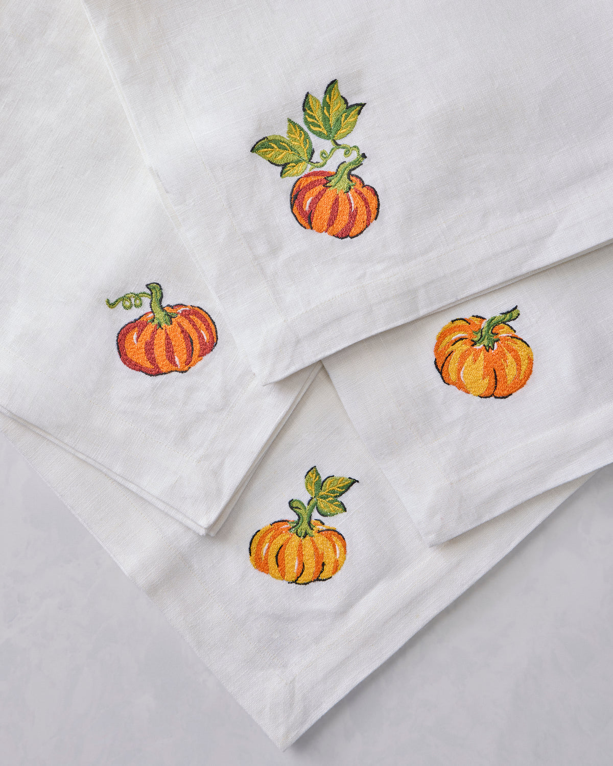 Close-up of four white dinner napkins overlapping, each with an embroidered orange pumpkin and green leaves.