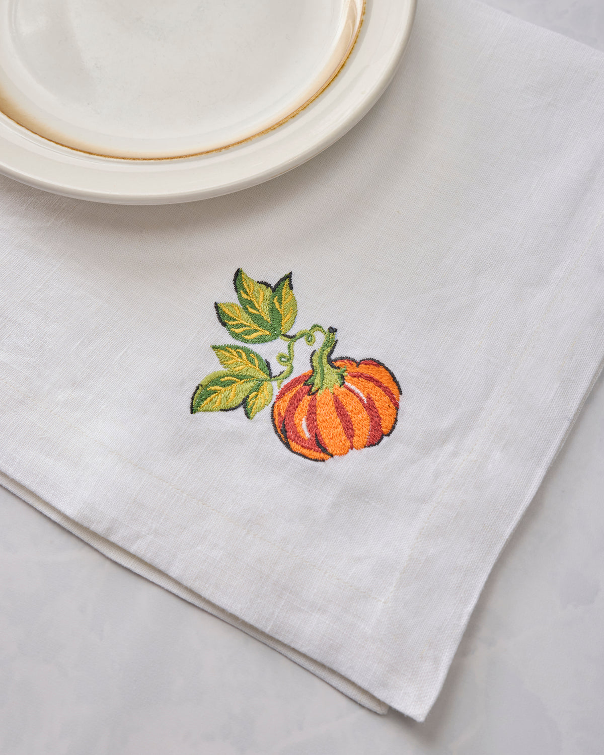 Close-up of a white dinner napkin with an embroidered orange pumpkin and green leaves beside stacked plates.