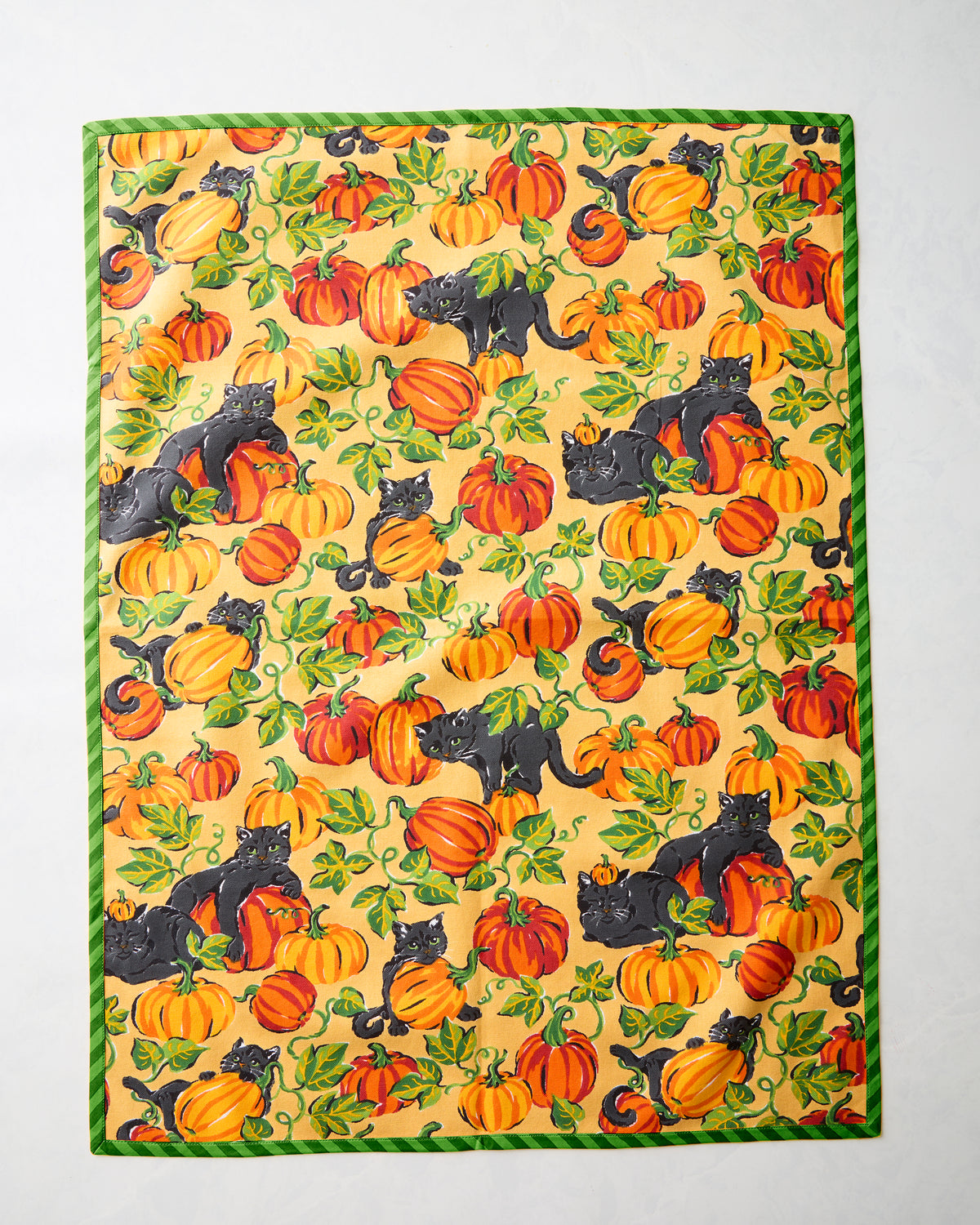 Tea towel with a repeating pattern of black cats, pumpkins, leaves, and vines, bordered in green.