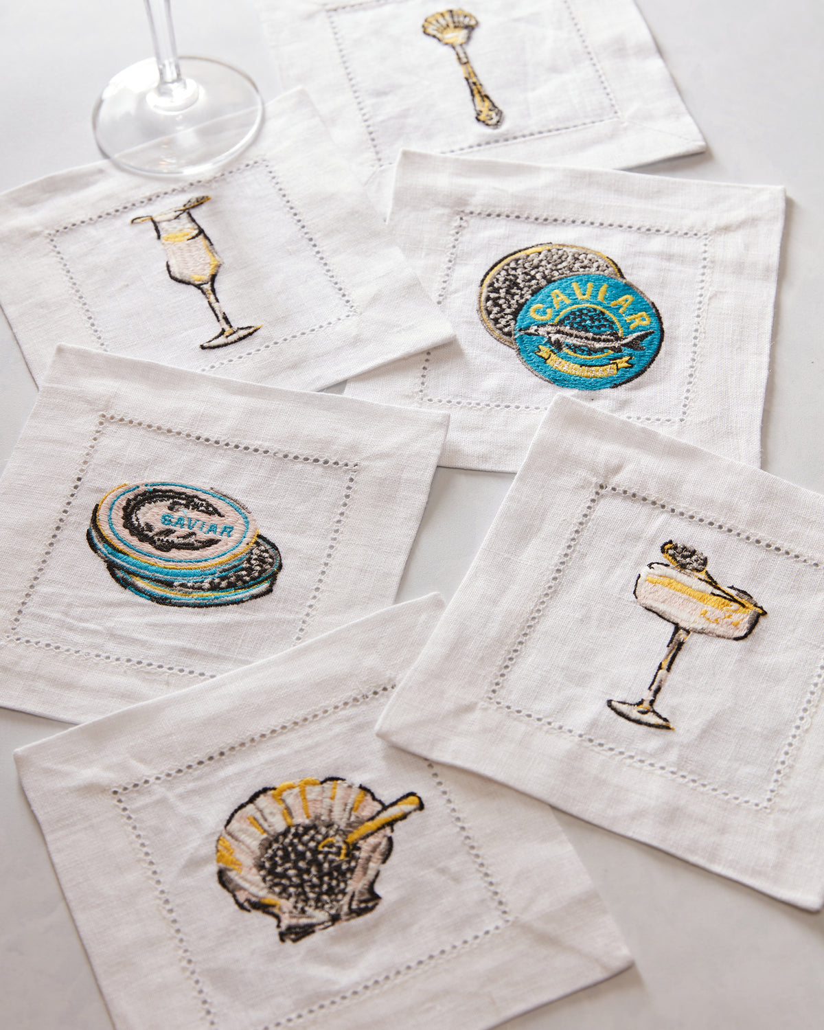 Several white Champagne & Caviar cocktail napkins arranged on a surface, embroidered with caviar tins, caviar, and champagne glasses; a wine glass is nearby.