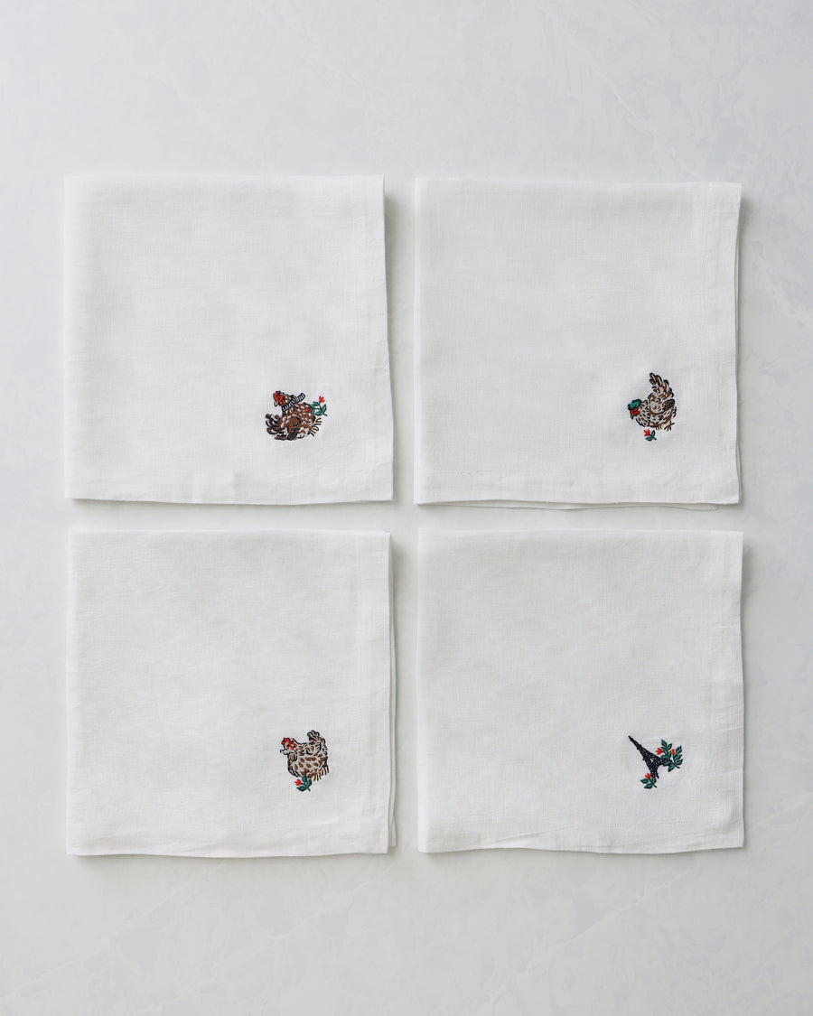 Four white dinner napkins arranged in a square, each with a small embroidered holiday motif near one corner.