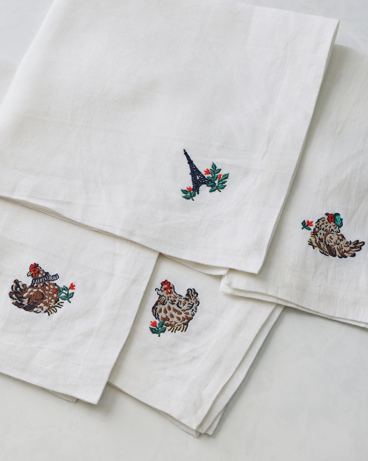 Close-up of four overlapping white dinner napkins showing embroidered animal, bird, and foliage motifs.