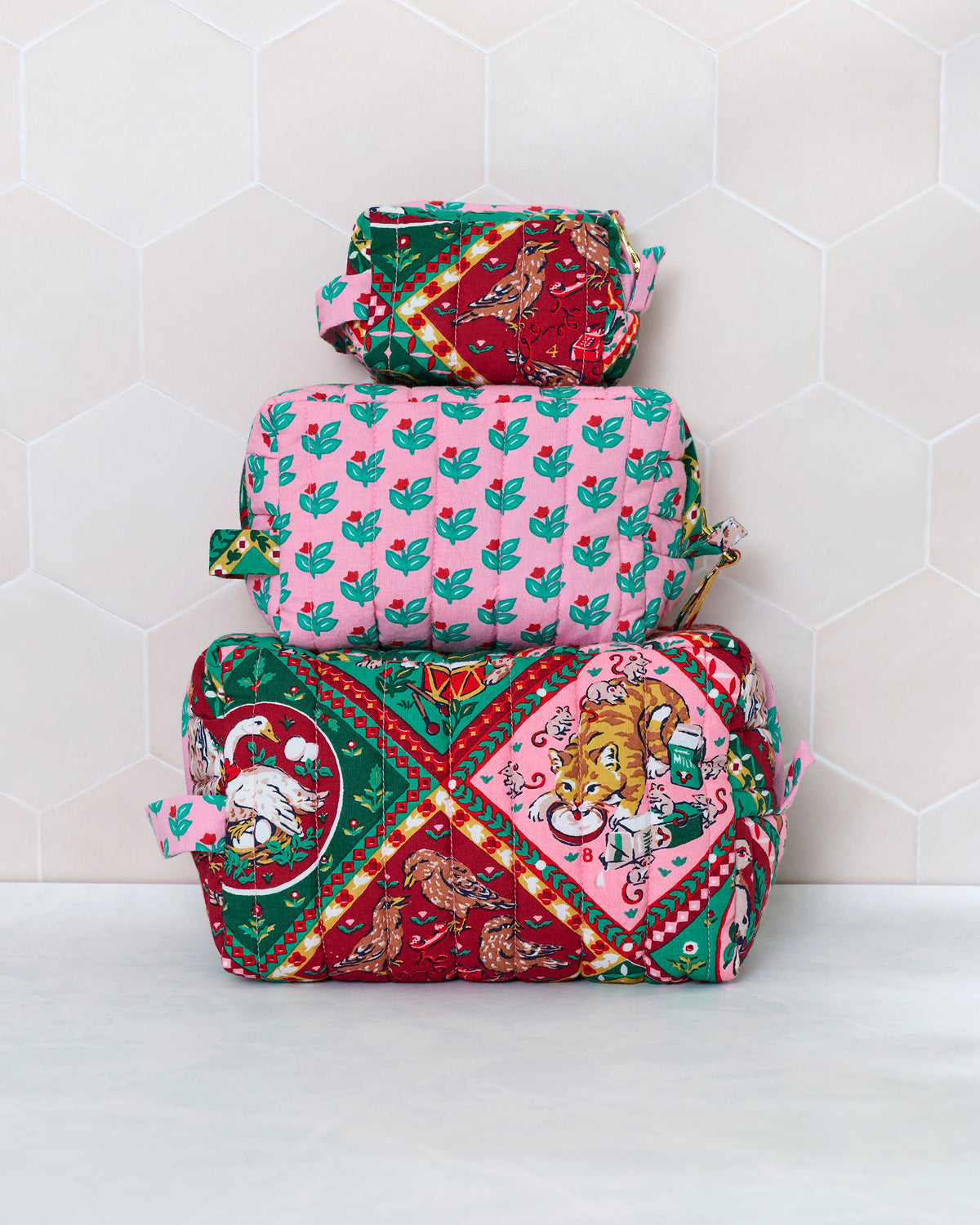 Three quilted 12 Days of Christmas pouches stacked by size, with the largest showing red, green, and pink holiday scenes and the middle pouch covered in a pink leaf print.
