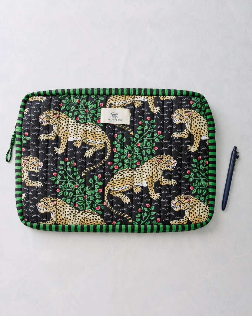 Black quilted laptop sleeve with green trim and leopard-and-leaf print, shown flat beside a pen.