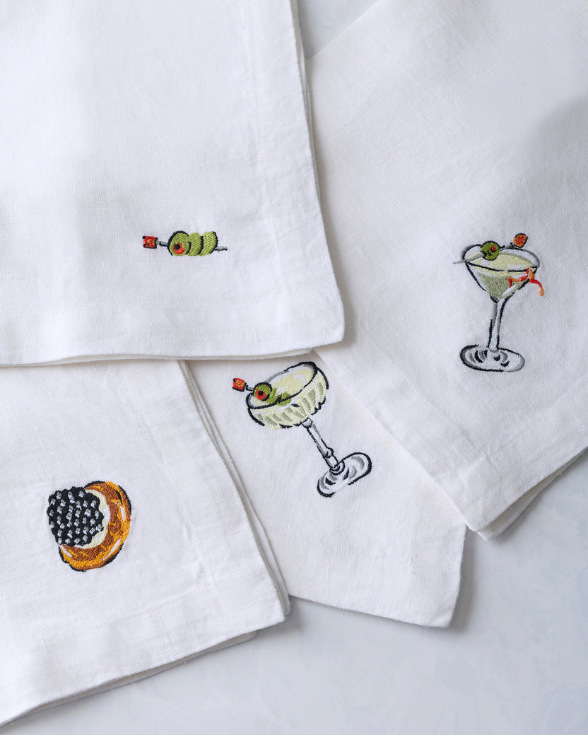 Close-up of four white dinner napkins embroidered with small food and cocktail motifs, including a caviar-topped canapé and cocktail glasses.