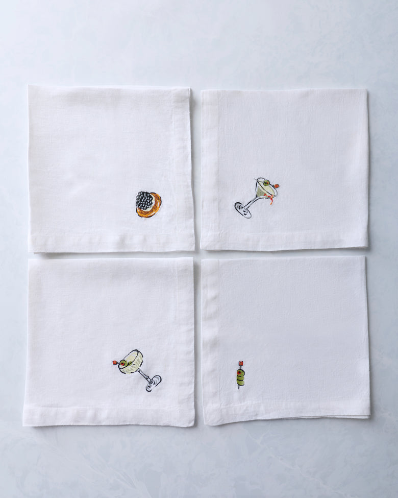 Four white dinner napkins arranged in a grid, each with a small embroidered food or cocktail motif.