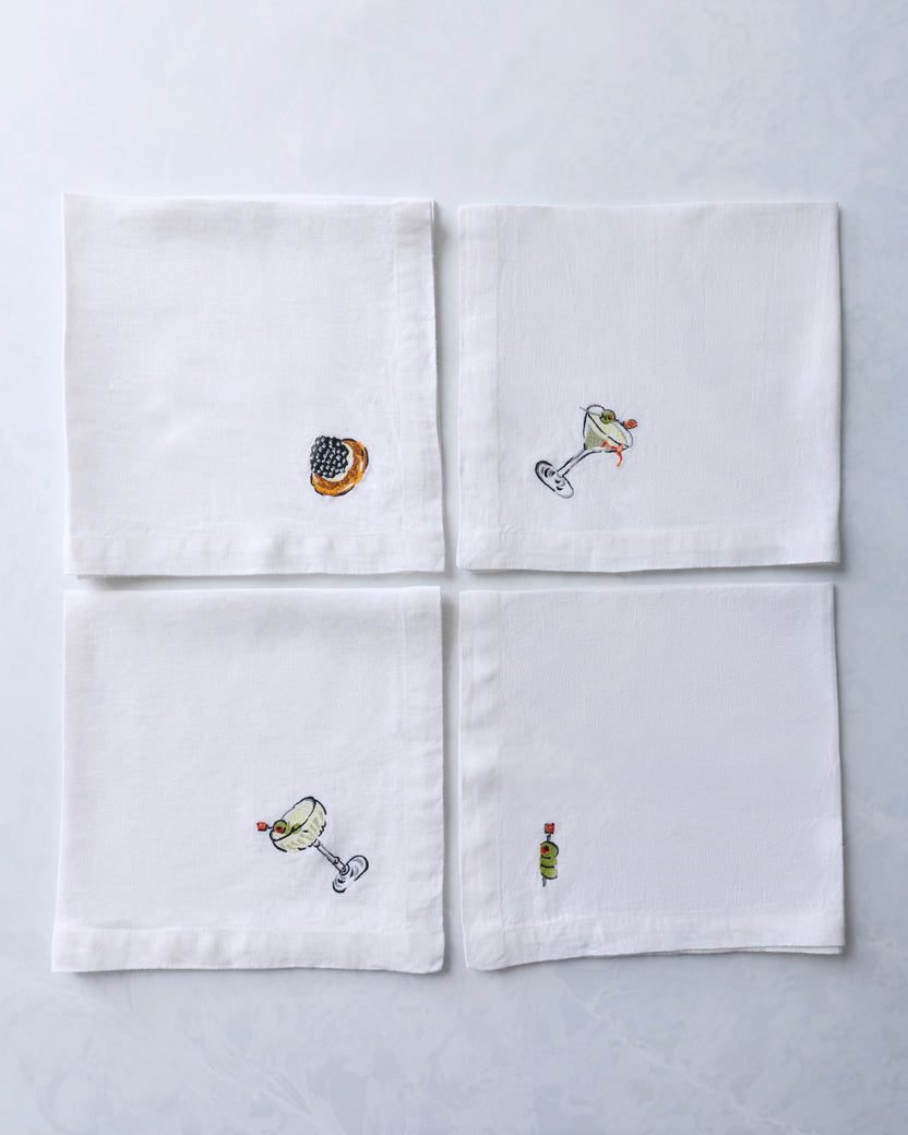 Four white dinner napkins arranged in a grid, each with a small embroidered food or cocktail motif.