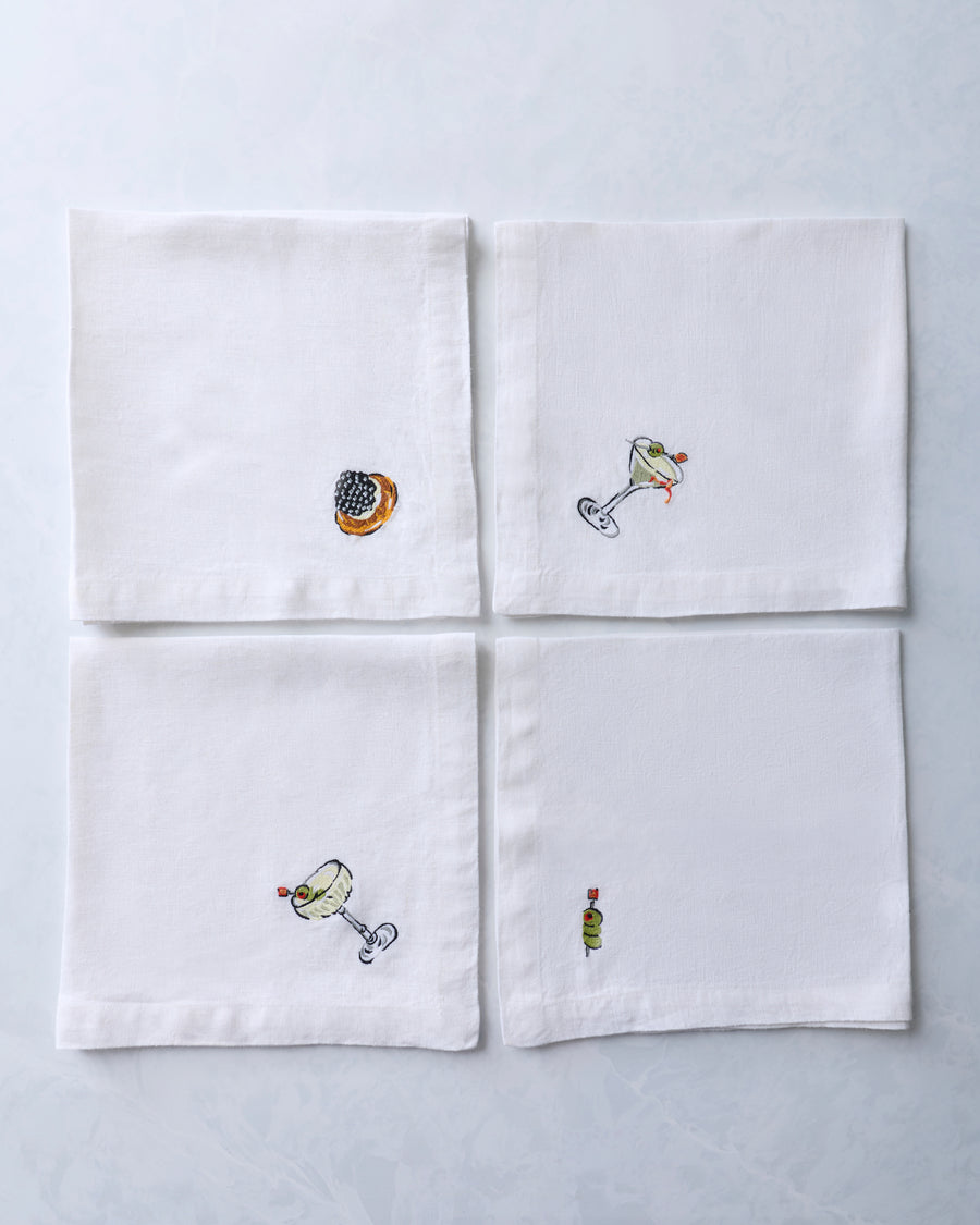 Four white dinner napkins arranged in a grid, each with a small embroidered food or cocktail motif.