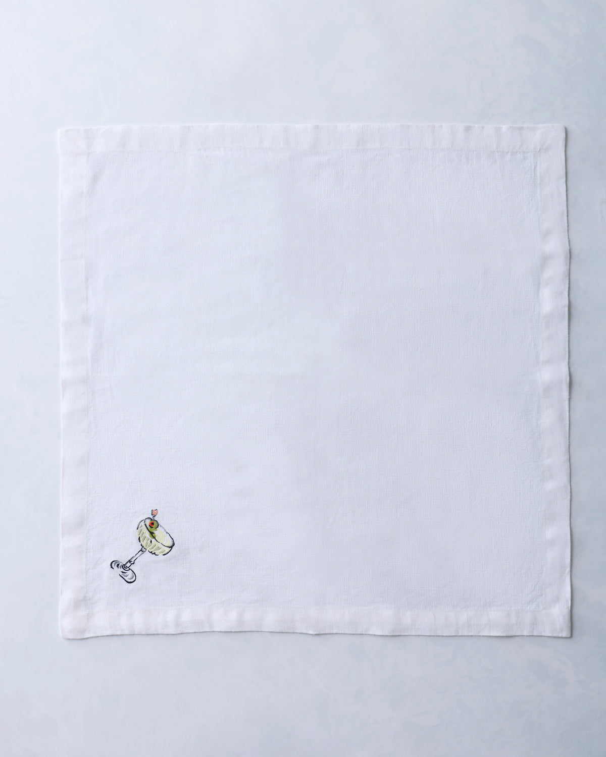 White square dinner napkin embroidered with a small cocktail glass and garnish motif near one corner.