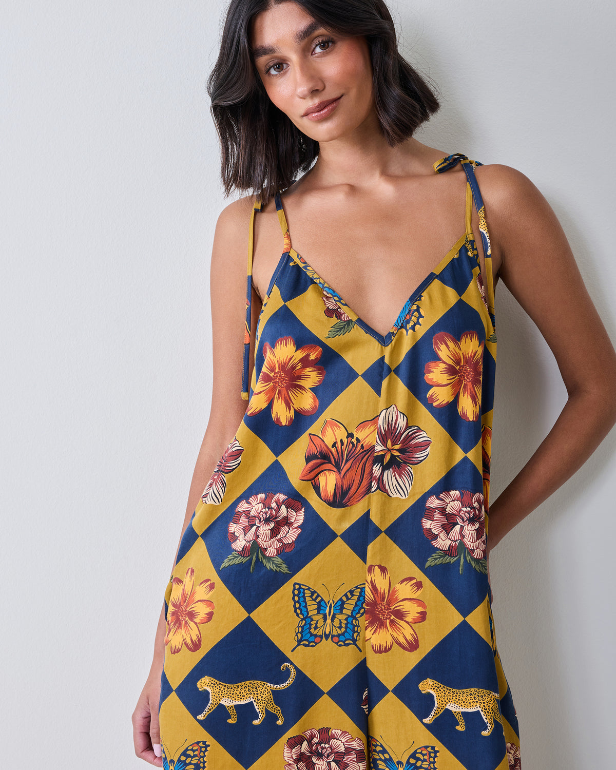 Front close-up of the Botanical Checkerboard Jaipur jumpsuit showing the V-neckline, tie shoulder straps, and floral, butterfly, and leopard motifs.