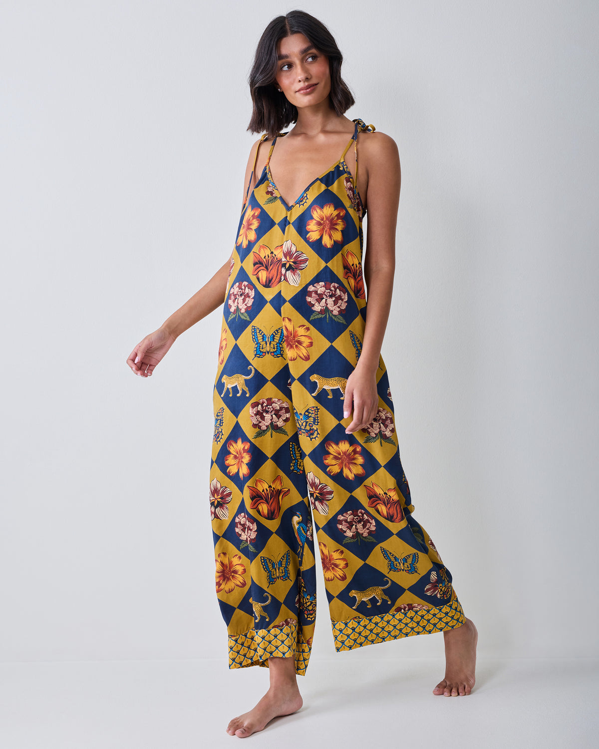 Front view of the Botanical Checkerboard Jaipur jumpsuit with tie shoulder straps, a V-neckline, wide legs, and floral, butterfly, and leopard motifs.