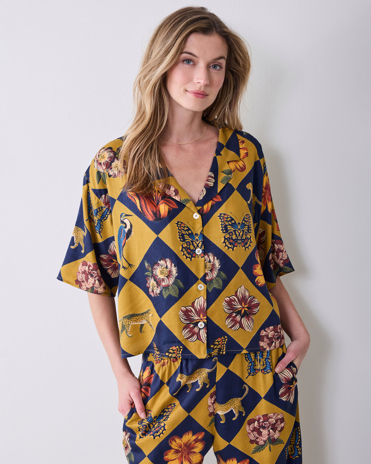 Front upper-body view of a person wearing the Golden Hour Botanical Checkerboard short-sleeved button-front top with floral, butterfly, and animal motifs.