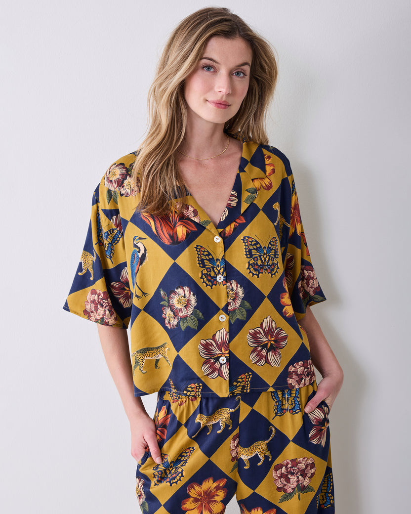 Front upper-body view of a person wearing the Golden Hour Botanical Checkerboard short-sleeved button-front top with floral, butterfly, and animal motifs.