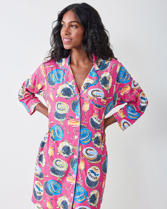A woman modeling a pink long-sleeve button-up shirt dress with a colorful graphic print of pomegranates, cocktail glasses, and citrus slices, set against a plain light gray background.